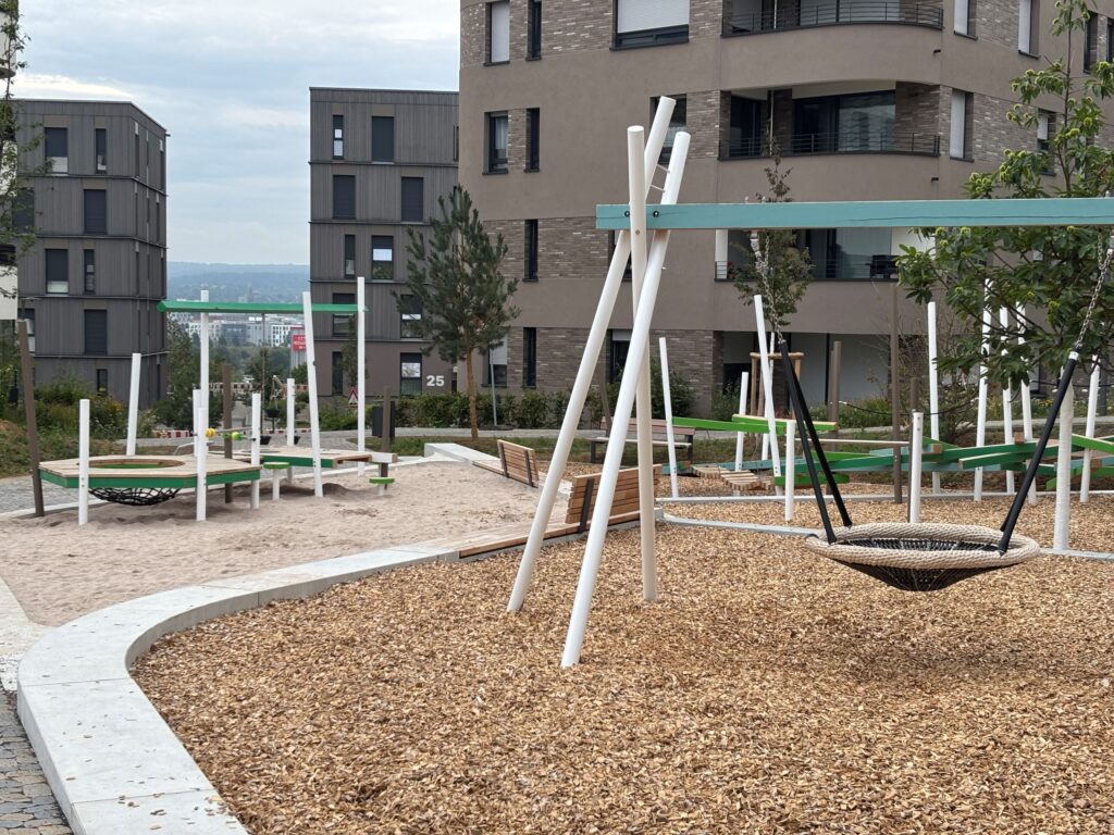 Spielplatzbereich mit Vogelnestschaukel, Sandkasten und Balanciergeräten vor den Wohngebäuden des Quartiers Hochgelegen